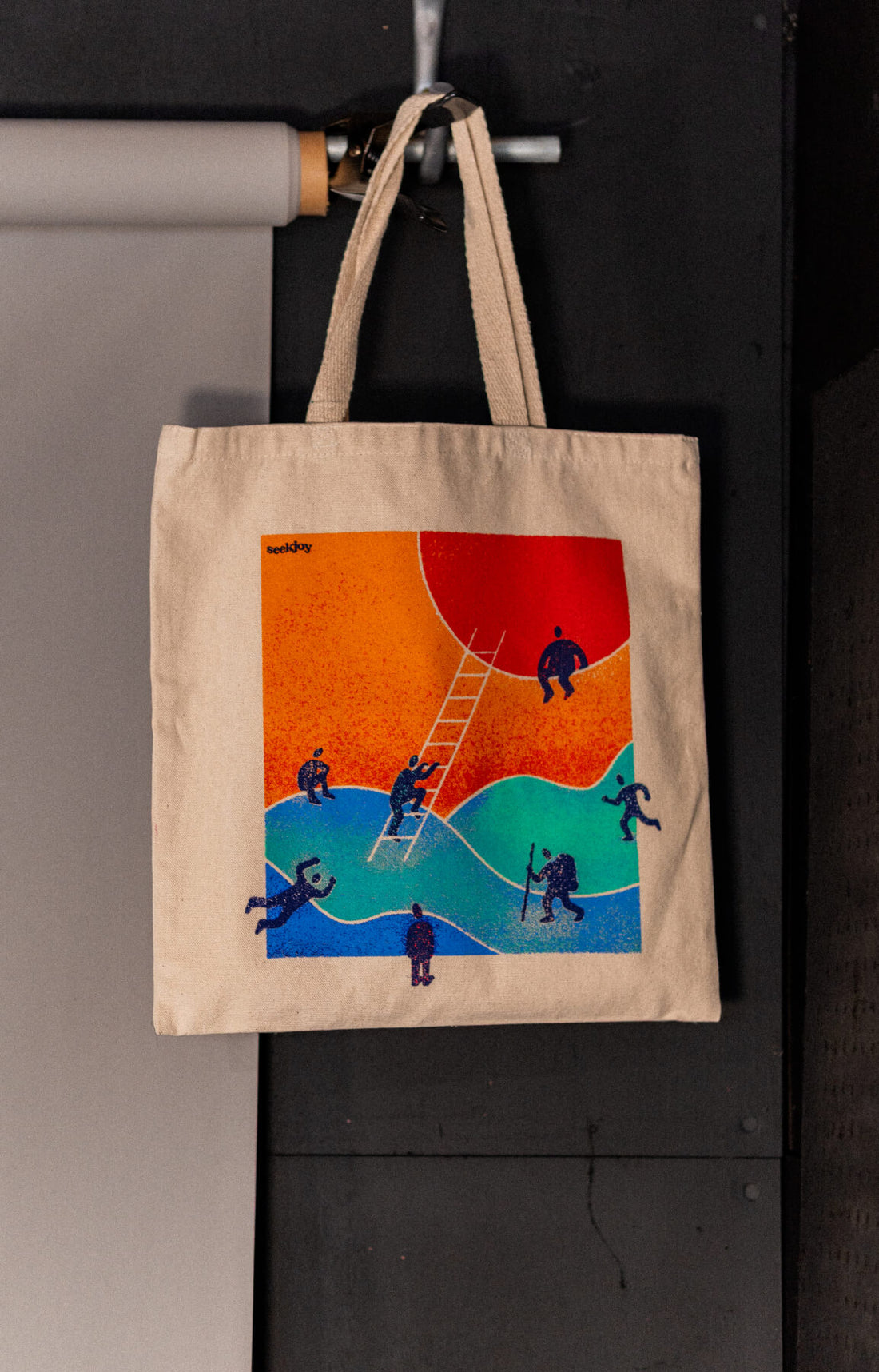 Aspirations tote hanging on a hook, orange and blue print of small figures climbing